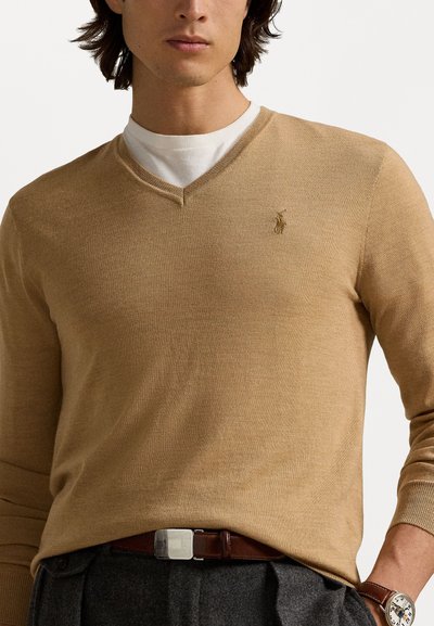 Pull beige à col en v en maille texturée, porté sur une chemise blanche, avec un petit logo sur la poitrine gauche.