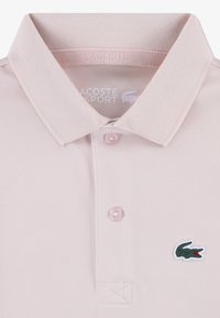 Light pink Lacoste polo shirt with a textured fabric, traditional collar, three buttons, and a decorative black and red crocodile logo.