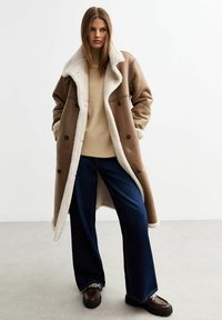 Abrigo de piel de oveja marrón con forro blanco, gran cuello con muescas y botones de doble botonadura, combinado con un suéter beige y jeans oscuros de pierna ancha.