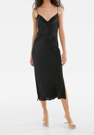 Kobieta w czarnej satynowej sukience midi na cienkich ramiączkach z bocznymi rozcięciami, zestawiona z przezroczystymi sandałami na obcasie.