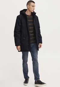 Modra parka jakna s kapuco, sprednjimi žepe in gumbnim zapiranjem. Nosimo jo preko prugaste majice in v kombinaciji z modrimi jeans hlačami ter črnimi supergami.