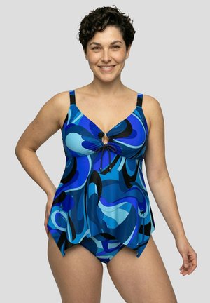 Femme aux cheveux courts et bouclés portant un maillot tankini à motif abstrait bleu et noir, souriante, debout devant un fond gris.