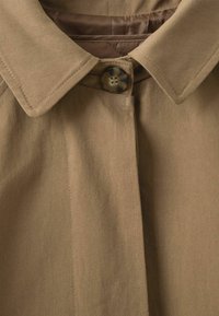 Beige külső öltözet gombos záródással, hegyes gallérral és finom átlós mintázattal. Simag belső bélés, masszív varrási részletek.