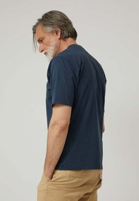 Hombre mayor con cabello y barba canosos, vistiendo una camiseta azul marino y pantalones beige, de pie de lado con las manos en los bolsillos.