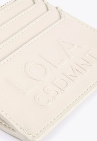 Närbild av en ljusbeige läderplånbok med präglingen "LOLA CSDMNFT" och synlig sömnad längs kanterna och kortfacken.