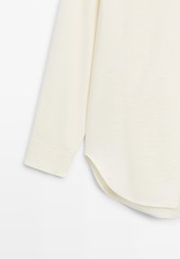 Camicia a maniche lunghe color crema chiaro con orlo arrotondato, tessuto morbido e polsini aderenti. Il tessuto sembra leggermente strutturato e leggero.