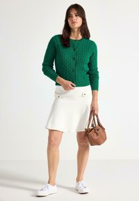 Grön kabelstickad cardigan med rund hals och knappar, i kombination med en vit A-linjeformad kjol och brun handväska; vita sneakers fulländar looken.