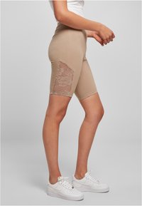 Beige fietsshorts met een kanten paneel aan de zijkant. Gladde stof, aansluitend design en midden-dijlengte. Gepaard met witte sneakers.