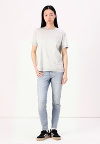 Camiseta de algodón gris claro con mangas cortas, combinada con jeans ajustados azul claro y zapatillas negras con rayas blancas y suelas marrones.