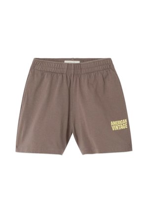 Lyshåret barn stående iført en mørkeblå hættetrøje, brune shorts og brune flip-flops mod en ensfarvet lys baggrund.