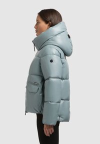 Chaqueta acolchada tipo puffer en azul claro, con un alto cuello, capucha de gran tamaño, bolsillo delantero y acentos de hardware negro. Textura suave y brillante.