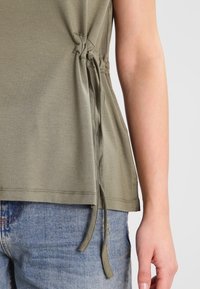 Olivgrön linne med sidoband för justerbar passform, gjort av mjukt tyg, med en avslappnad passform, bärs över ljusblå denimjeans.