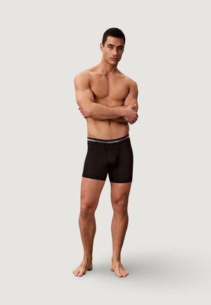 Gespierde man met kort haar, blootsvoets staand, met gekruiste armen, dragend zwarte Calvin Klein boxershorts tegen een effen lichte achtergrond.
