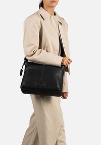 Borsa a spalla nera in pelle con una superficie liscia, forma rettangolare e un sottile accento con il logo sul davanti. Indossata sopra un giubbotto beige.