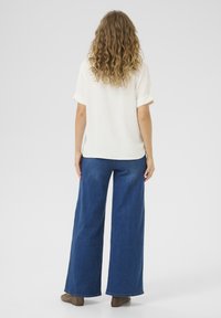 Camisa blanca de manga corta con puños enrollados, combinada con pantalones de mezclilla anchos de color azul y botas de tobillo marrones, vista desde la parte posterior.