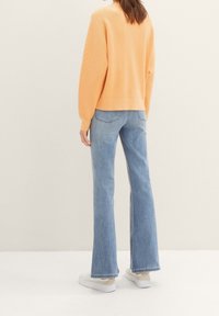 Pomarańczowy prążkowany sweter w połączeniu z jasnoniebieskimi dzwonowatymi dżinsami i białymi tenisówkami, widziane od tyłu na neutralnym tle.