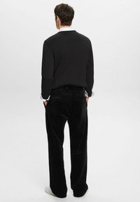 Musta pitkähihaine paita valkoisella kauluksella, yhdistettynä mustiin korduroy-housuihin. Malli kääntyi selkä menosuuntaan, jalassaan valkoiset lenkkitossut. Yksinkertainen muotoilu.