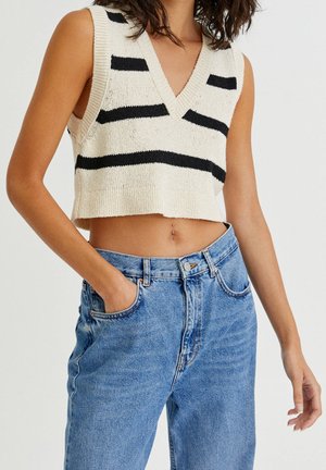 Gilet tricoté court sans manches à rayures noires, porté avec un jean en denim bleu taille haute, une main dans la poche, ventre découvert.