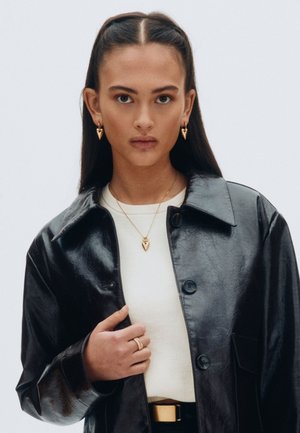 Schwarze Kunstlederjacke mit Kragen, darunter ein weißer Pullover, goldene herzförmige Ohrringe und Anhänger sowie ein Ring an der Hand.