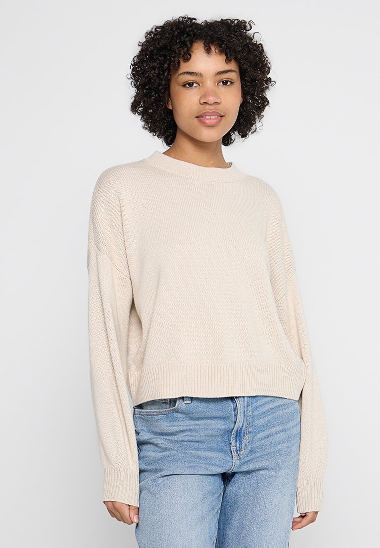 Maglione beige corto con lavorazione a maglia testurizzata, scollo rotondo e spalle cadenti, abbinato a jeans blu a vita alta.