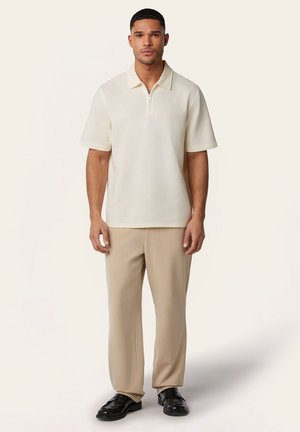 Man staat en draagt een crèmekleurige korte mouw polo met rits, beige broek en zwarte loafers tegen een effen lichte achtergrond.