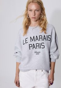 Ung kvinna med blont hår bär en grå sweatshirt med texten "Le Marais Paris France 75003" och vita byxor, står mot en enfärgad bakgrund.