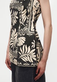 Gros plan d'un haut noir sans manches avec des motifs botaniques et géométriques beiges, porté avec un pantalon foncé.