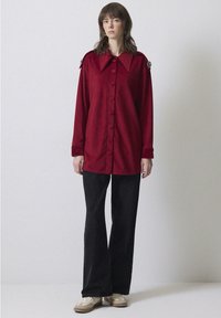 Camicia di velluto a coste rossa con un ampio colletto a punta, chiusura con bottoni e maniche lunghe, abbinata a pantaloni neri a zampa d'elefante e sneaker beige.