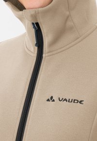 Beige Fleecejacke mit hohem Kragen und schwarzem Reißverschluss. Verfügt über ein dezentes Logo auf der Brust und eine glatte Textur.
