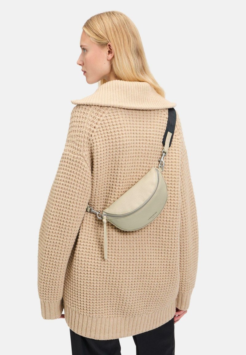 Pull en tricot beige surdimensionné avec un grand col, associé à un petit sac en cuir vert clair en bandoulière, courbé, avec une fermeture éclair en argent.
