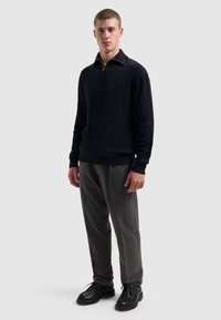 Een jonge man staat in een zwart half-rits sweater, grijze broek en zwarte veterschoenen tegen een effen lichte achtergrond.