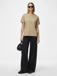 Žena koja nosi sunčane naočale sa okvirom u boji kornjačevine, bež majicu, crne hlače širokih nogavica, crne cipele i drži malu crnu torbicu.