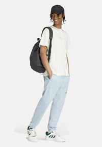 Camiseta blanca de manga corta con el logo de Adidas, pantalones jogger azul claro, mochila negra y zapatillas con detalles verdes; llevando una gorra negra.