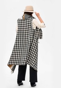 Czarnobiała chusta w kratę z frędzlami, nałożona na kremowy top, noszona z czarnymi spodniami i beżowym kapeluszem.
