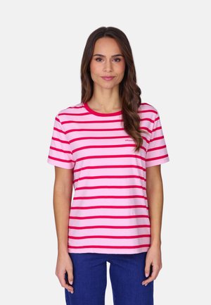 Femme aux longs cheveux bruns portant un t-shirt à manches courtes rayé rose et rouge et un pantalon bleu, debout devant un fond uni.