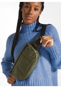 Borsa a spalla verde oliva trapuntata con chiusura zip, tracolla nera e dettaglio logo embrittato. Indossata da un modello con un maglione blu.