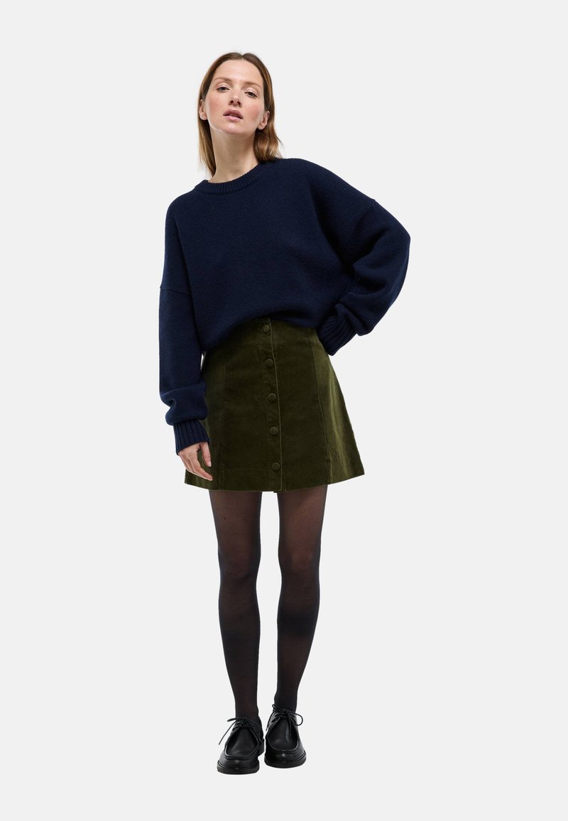 Femme debout devant un fond blanc, portant un pull oversize bleu marine, une jupe boutonnée devant couleur vert olive, des collants noirs et des chaussures noires.
