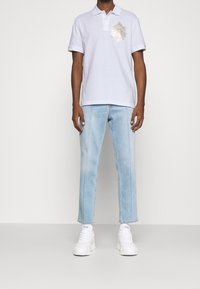 Polo-Shirt in Hellgrau mit einem dekorativen goldenen Emblem auf der Brust, kombiniert mit hellblauen geraden Jeans und weißen Sneakern.