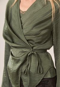 Top envolvente de color verde oliva, hecho de una tela ligera y texturizada. Presenta un lazo en la parte delantera y un diseño en capas con mangas largas y un dobladillo asimétrico.
