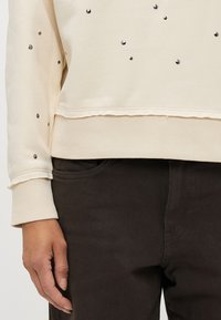 Ανοιχτό μπεζ φούτερ με μεταλλικά τρουκς; ριγμένα μανίκια και στρίφωμα; συνδυασμένο με σκούρα καφέ παντελόνια. Μαλακή υφή και άνετη γραμμή.