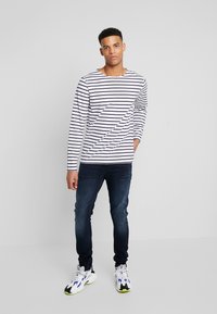 Camicia a maniche lunghe a righe bianca e blu navy, dal design aderente, abbinata a jeans scuri e sneakers bianche con accenti colorati.