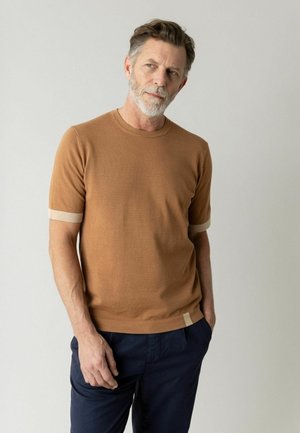 Muž so sivou bradou, oblečený v hnedej košeli s krátkym rukávom a béžovými ozdobami na manžetách, a tmavomodrých nohaviciach, stojaci s jednou rukou v vrecku.