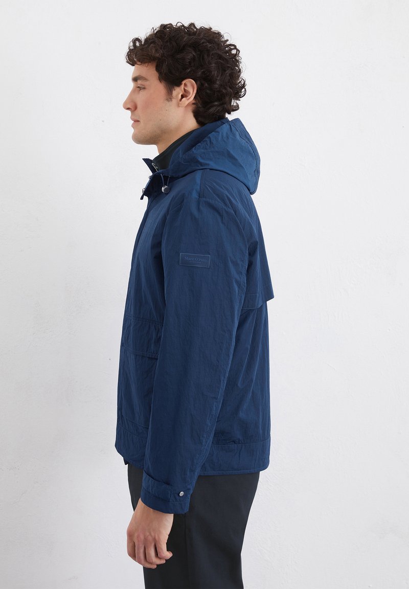 Marineblauwe lichtgewicht jas met een capuchon, zijzakken en drukknoopsluitingen. Heeft een geweven stof en een label op de mouw.