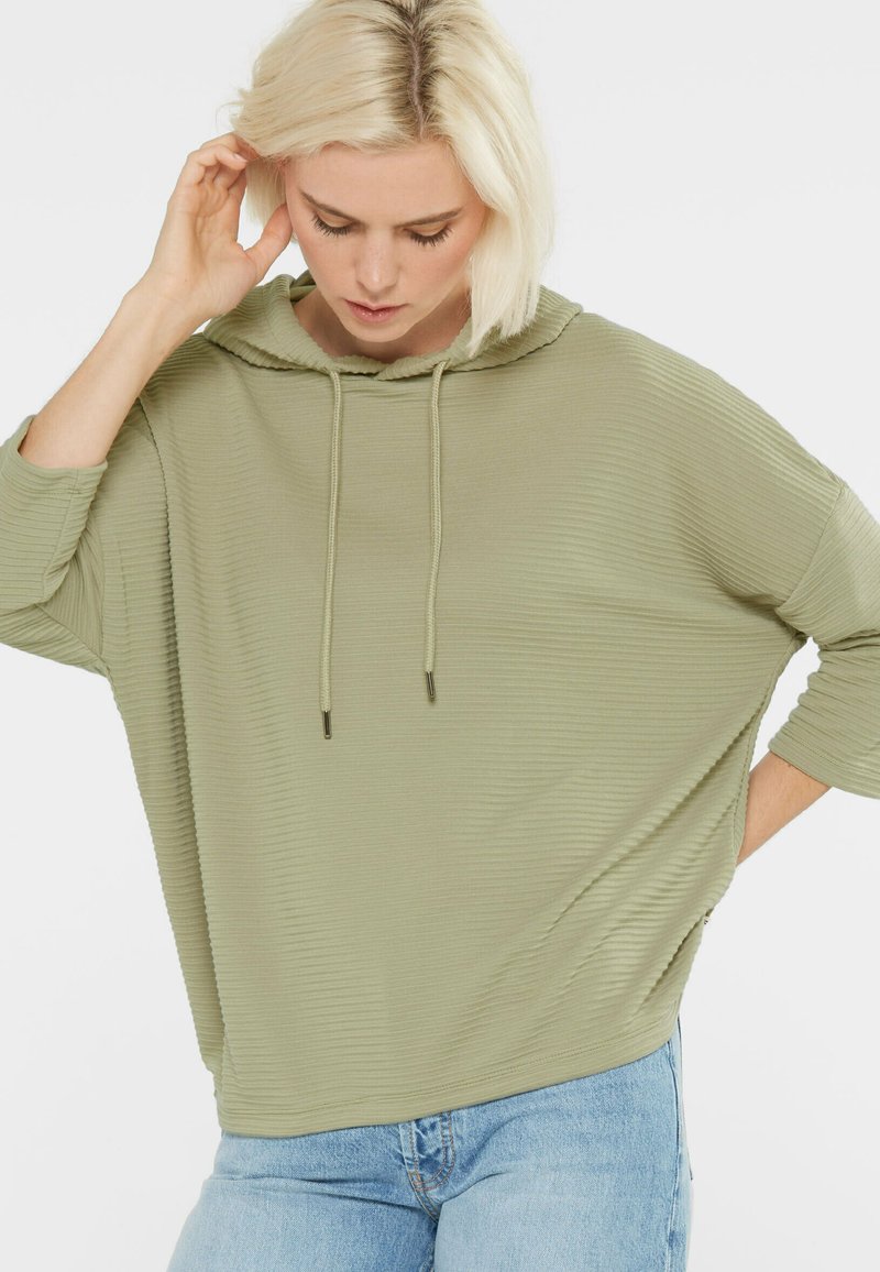 Olive green hooded sweatshirt with a ribbed texture, loose fit, and drawstring. Worn with light blue denim jeans.