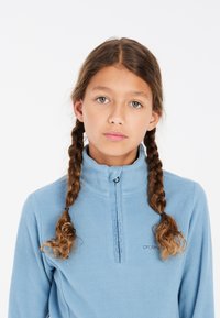 Pullover in pile azzurro chiaro con chiusura a mezza zip, colletto alto e dettaglio del logo. Presenta un tessuto testurizzato e trecce che incorniciano il viso.