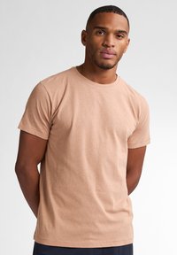 Světle hnědé bavlněné tričko s krátkým rukávem, kulatým výstřihem a hladkým povrchem. Jednoduchý design, nošené s rukama za zády.