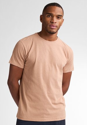 T-shirt di cotone marrone chiaro con maniche corte, colletto tondo e una texture liscia. Design semplice, indossata con le mani dietro la schiena.
