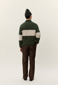 Férfi áll zsebébe tett kézzel, zöld és bézs színű hosszú ujjú felsőt, barna nadrágot, fekete cipőt és zöld sapkát visel, háttal a nézőnek.