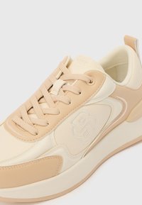 Bež sintetička tenisica s teksturiranim završetkom, vezicama sprijeda i diskretnim logotipom sa strane, s kontrastnim kremastim potplatom.