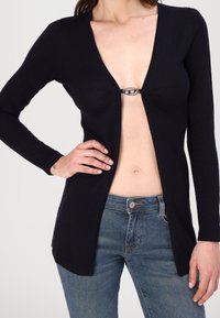 Chaleco negro de manga larga con un profundo escote en V y un detalle de broche en el frente. Combinado con unos jeans de denim azul con cierre de botón.